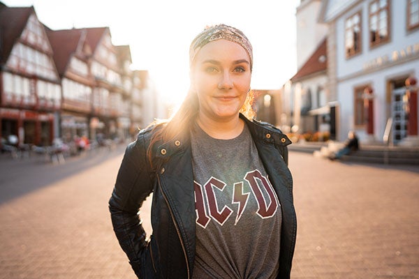 Eine junge Frau steht mitten in einer Kleinstadt und lächelt in die Kamera.