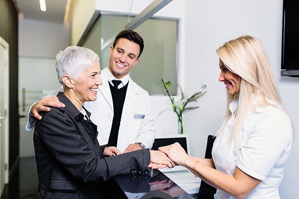 Ein Arzt und eine Therapeutin begrüßen eine Patientin herzlich und halten ihre Hand. 