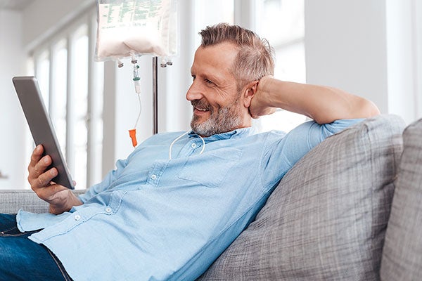 Ein Mann liegt mit einem Tablet auf der Couch, während er eine medizinische Infusion bekommt.