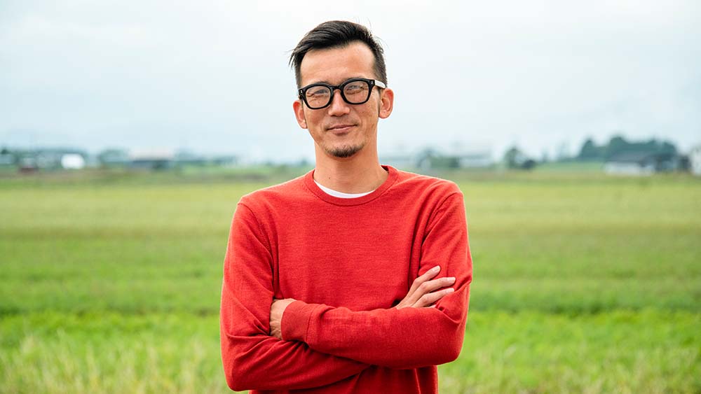 Eine Person mit Brille steht selbstbewusst und mit verschränkten Armen auf einem weiten Feld.